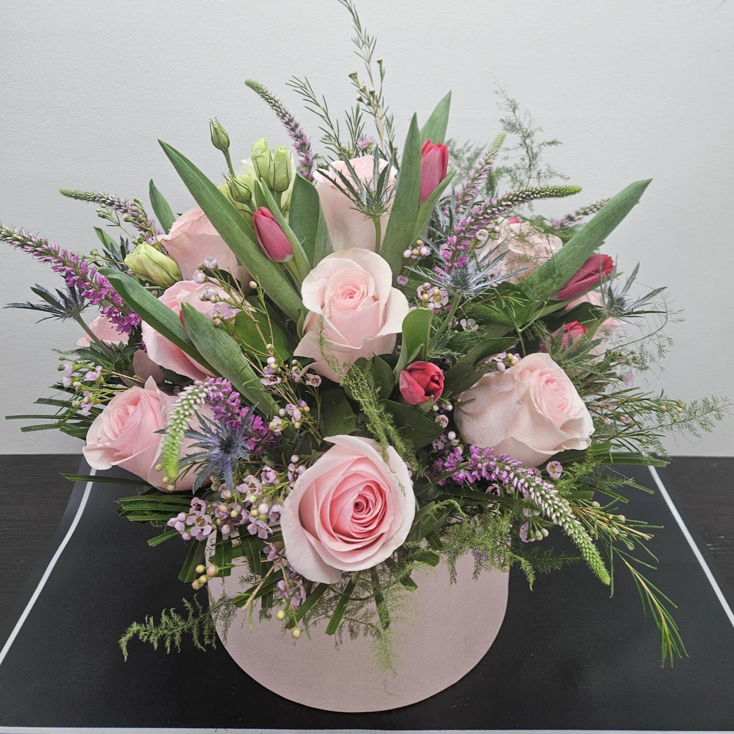 A floral arrangement featuring pink roses, tulips, and various small flowers in shades of pink and green, displayed in a round container.